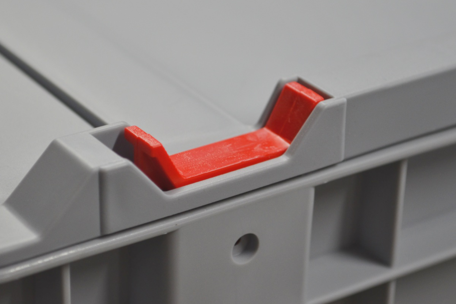 A gray storage container features a prominent red locking mechanism on its lid. The component aids in securing the lid in place, ensuring stability and protection of stored items. The background is neutral and unobtrusive.