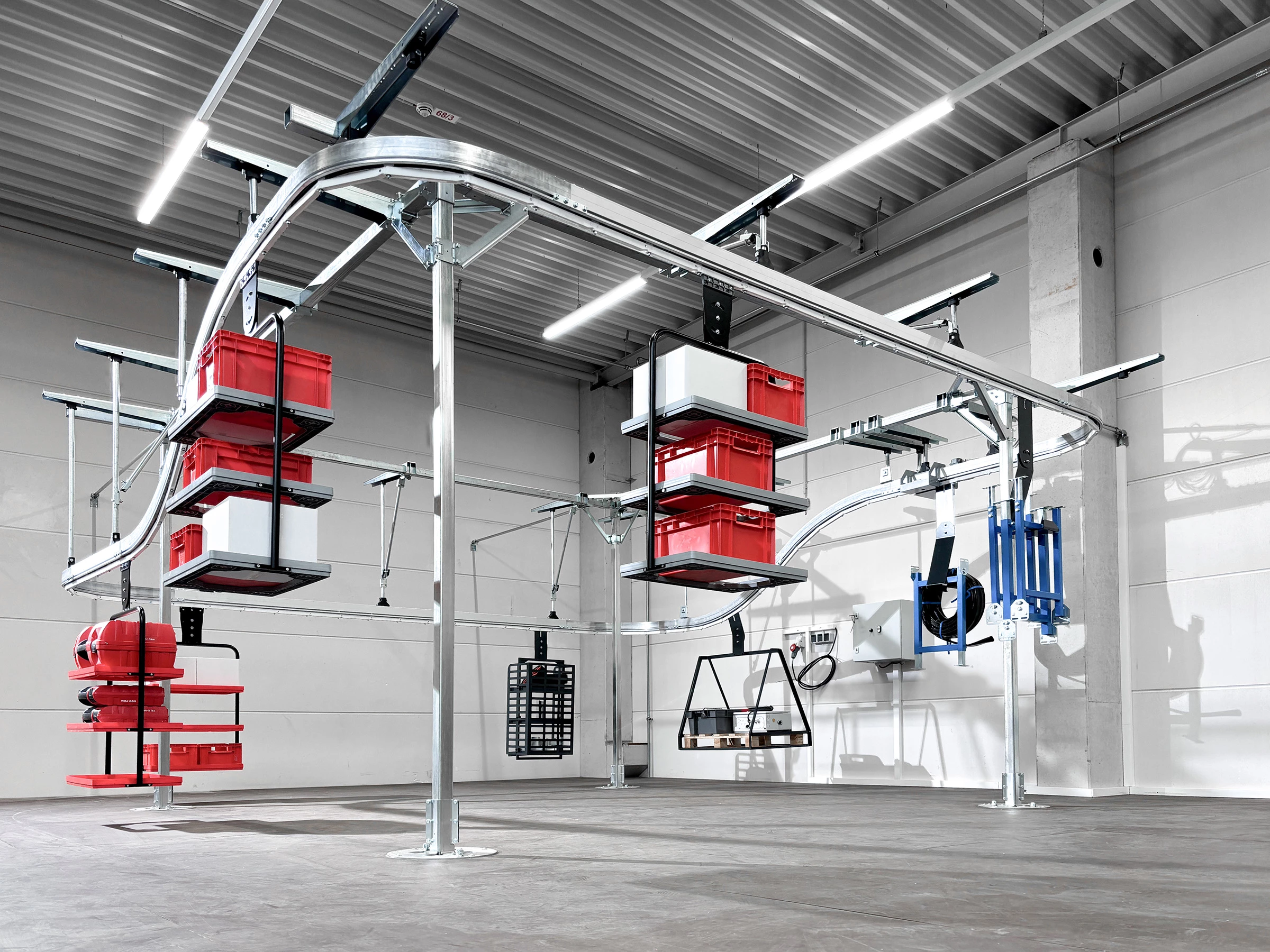 Hängendes Transportsystem mit roten und weißen Behältern, die auf Kurven und geraden Schienen platziert sind. Das System ist in einem modernen, beleuchteten Industrieumfeld installiert.