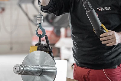 Ein Arbeiter hält eine schwere Metallkomponente mit einem Haken und einer Kette in einer Hand, während er mit der anderen Hand ein Werkzeug bedient. Im Hintergrund sind industrielle Bedingungen sichtbar.
