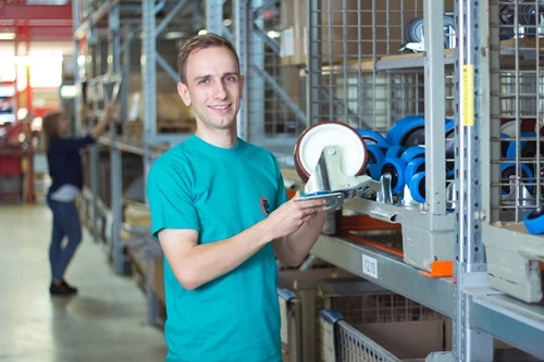 Ein junger Mann hält eine industrielle Rolle in der Hand. Er steht vor einem Regal mit verschiedenen Rädern und Rollen, während eine andere Person im Hintergrund beschäftigt ist. Der Raum wirkt hell und strukturiert.