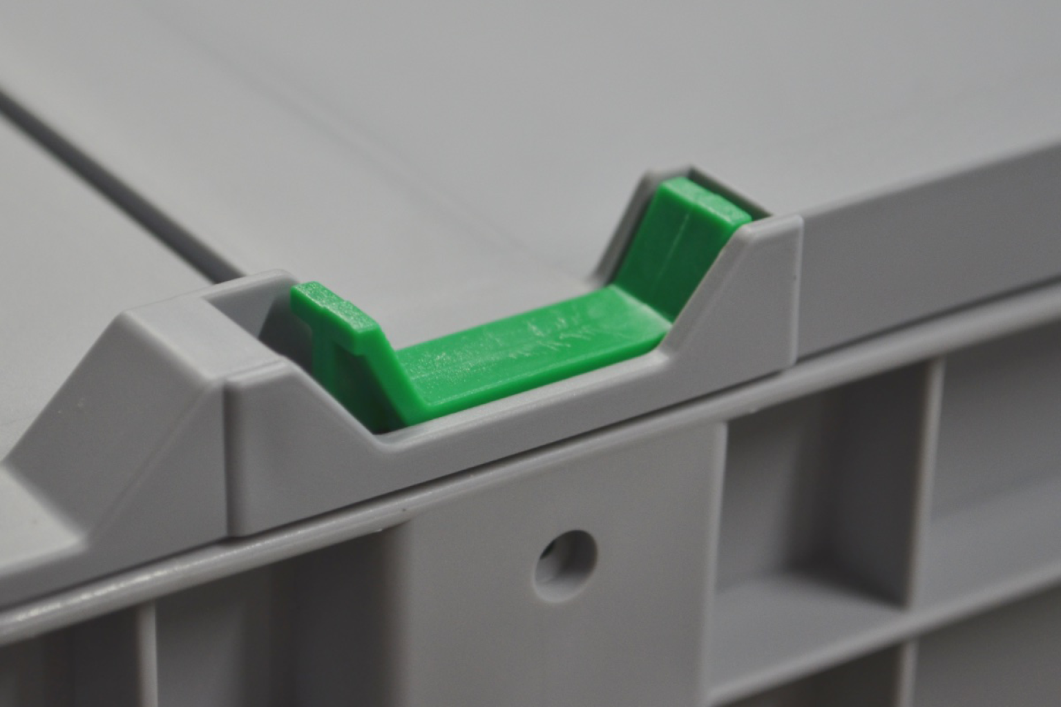 A gray plastic container features a green plastic latch or stopper located on the edge. The latch appears designed to secure the lid, ensuring stability during storage or transport. The background is blurred, emphasizing the container-s details.