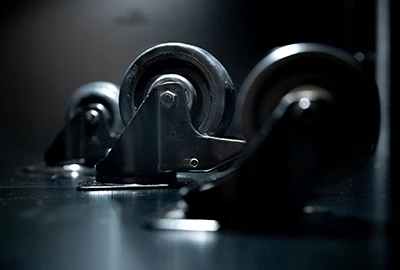 Drei industrielle Rollen sind nebeneinander auf einer dunklen Oberfläche angeordnet. Jede Rolle hat eine Metallbasis und gummierte Räder, die glänzend reflektieren. Die Umgebung ist stilistisch neutral.
