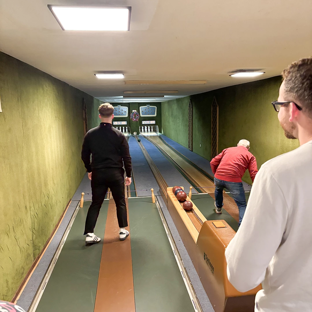 Zwei Personen befinden sich in einer Kegelbahn mit grünem Wandbelag. Einer steht in spielerischer Position, während der andere eine Kugel wirft. Im Hintergrund sind Kegel sichtbar.