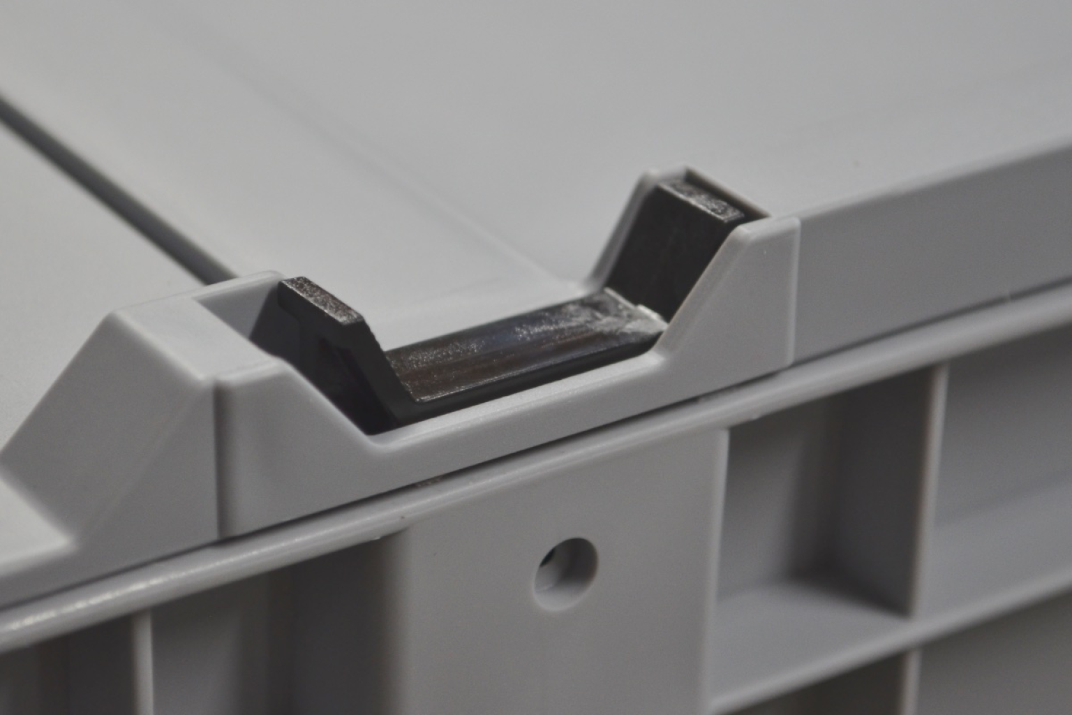 A grey plastic storage container is shown, focusing on a corner where the lid meets the body. The lid features a small black metal latch for secure fastening. The background is neutral to emphasize the container-s details.