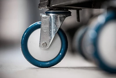 Ein blauer Gummi-Rad mit einer weißem Lauffläche ist an einer Metallachse montiert, platziert unter einem Transportwagen. Der Hintergrund zeigt mehrere ähnliche Räder.