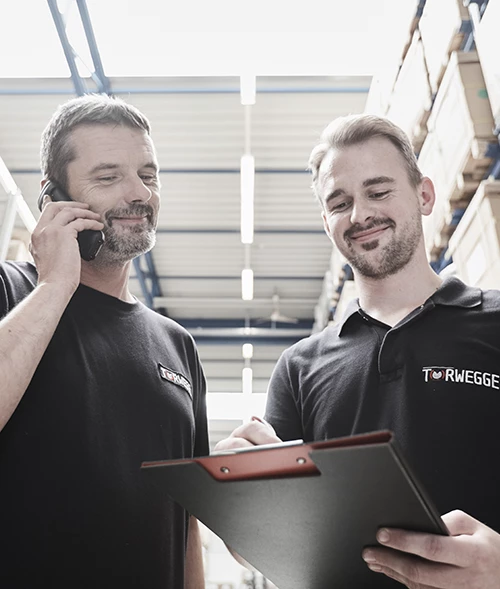 Zwei Männer in schwarzen Poloshirts stehen in einem Lager. Einer telefoniert, während der andere auf ein Clipboard schaut und Notizen macht. Regale mit Materialien sind im Hintergrund sichtbar.