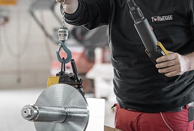 Ein Mitarbeiter hält einen großen Metallzylinder mit einem Haken an einer Kette in der linken Hand und einer elektrischen Schraubenschlüssel in der rechten Hand. Der Hintergrund ist unscharf und zeigt eine industrielle Umgebung.