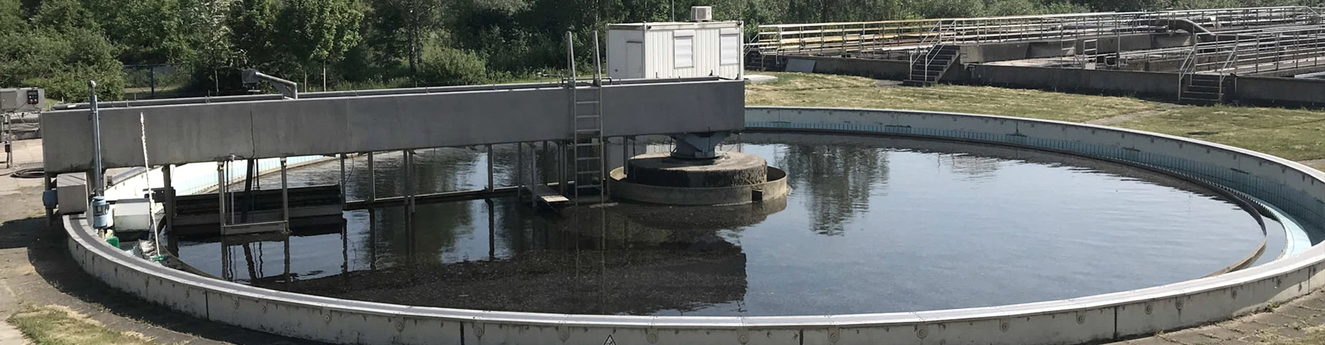 Ein kreisförmiges Becken mit klarem Wasser, in der Mitte eine rotierende Anlage zur Wasserreinigung. Im Hintergrund sieht man eine grüne Umgebung und einige industrielle Strukturen sowie eine Leiter.