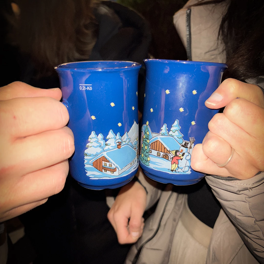 Zwei blaue Tassen mit festlicher Winterdekoration werden von zwei Händen gehalten. Die Tassen zeigen schneebedeckte Häuser, einen Weihnachtsbaum und eine Darstellung von Kindern, die einen Schneemann bauen.