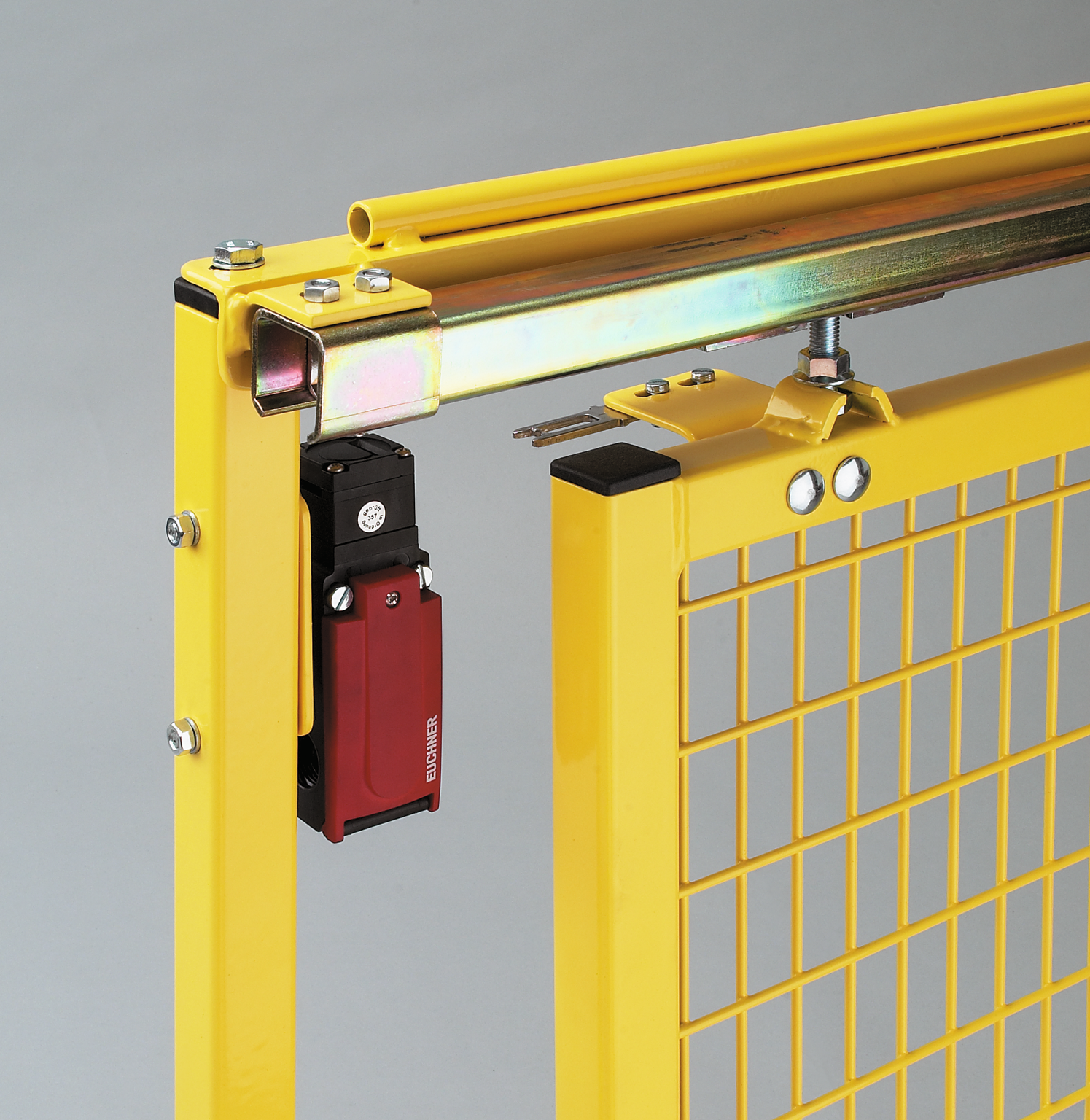 A yellow safety gate features a mesh design with a sliding mechanism, mounted on a rail. A red electronic switch is attached for operational control, positioned on the side. The background is a simple gray.