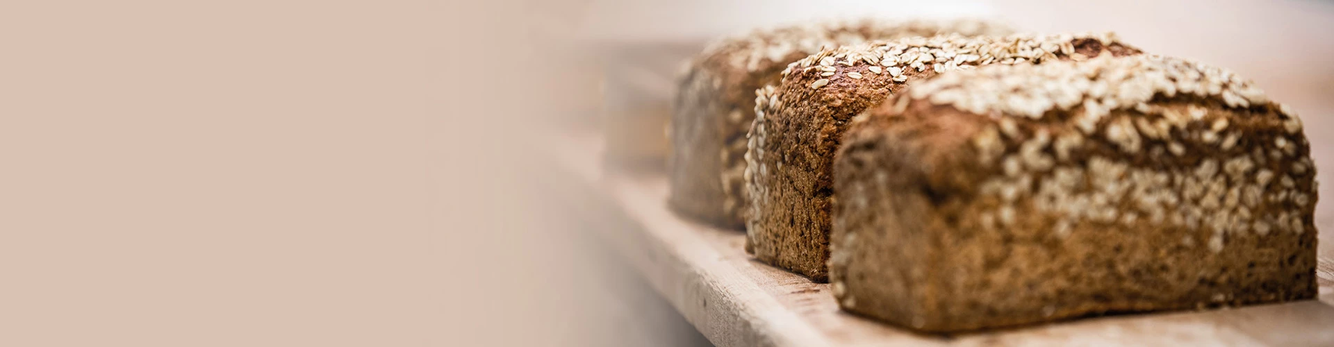 Drei frisch gebackene Laibe aus Vollkornbrot mit Haferflocken auf der Oberfläche liegen nebeneinander auf einem Holzbrett. Der Hintergrund ist unscharf und in sanften Farben gehalten.