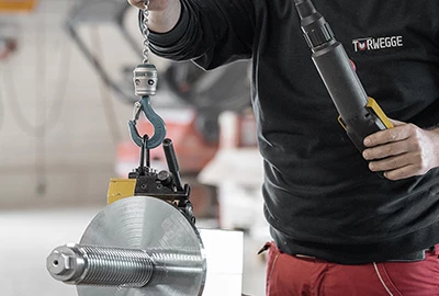 Ein Arbeiter hält eine große Metallrolle mit einer Vorrichtung in der Hand. In der anderen Hand hält er ein Aufnahmewerkzeug. Der Hintergrund zeigt eine industrielle Umgebung mit weiteren Werkzeugen.