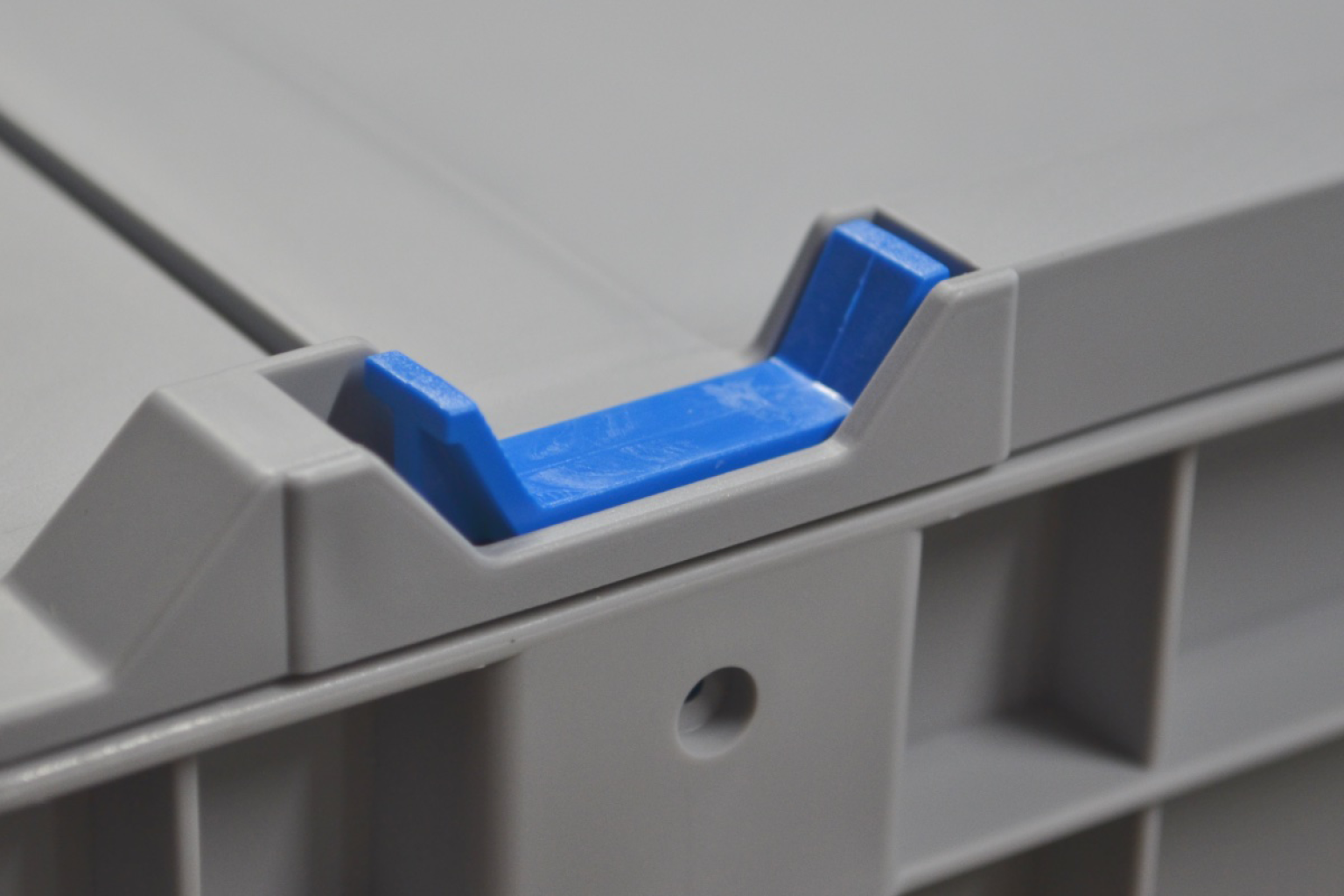 A gray plastic storage container features a blue locking mechanism integrated into the lid-s edge. The design facilitates secure closure in an industrial or warehouse setting.